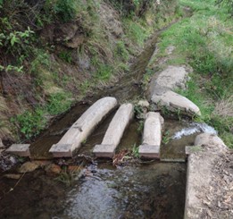 Sistema tradicional de reparto de agua en una acequia con compuertas de madera y piedra en un entorno rural.