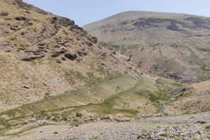 Vista panorámica de una ladera de alta montaña con vegetación escasa y senderos de agua o acequias integradas en el relieve rocoso.